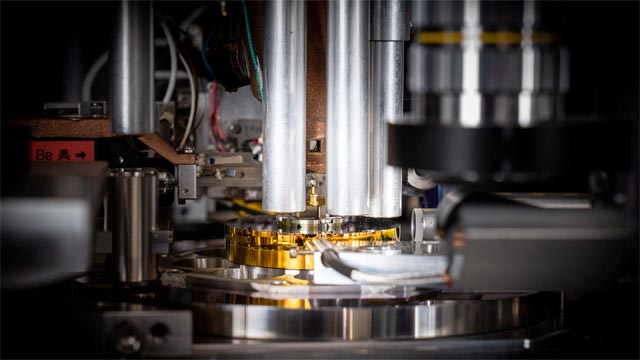 Die Probe, ein zylinderförmiger Ausschnitt aus einem handelsüblichen Computerchip, wird von der goldfarbenen Nadel in der Mitte des Bildes gehalten. Mit einem Durchmesser von weniger als 0,000 005 Meter (etwa 20 Mal kleiner als die Breite eines menschlichen Haares) wurde sie mit einem fokussierten Ionenstrahl aus dem Chip herausgeschnitten und auf dem Stift platziert. © Paul Scherrer Institut PSI/Mahir Dzambegovic